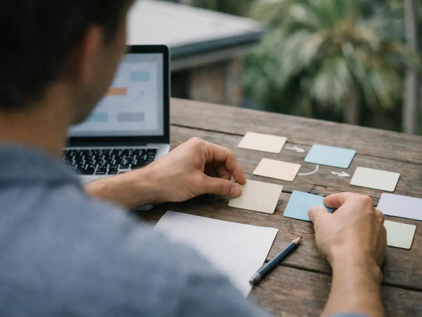 A person arranges colored cards on a wooden table beside a laptop outdoors