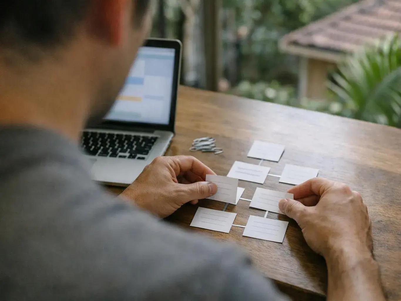 A person arranges small white cards on a wooden table beside an open laptop outdoors