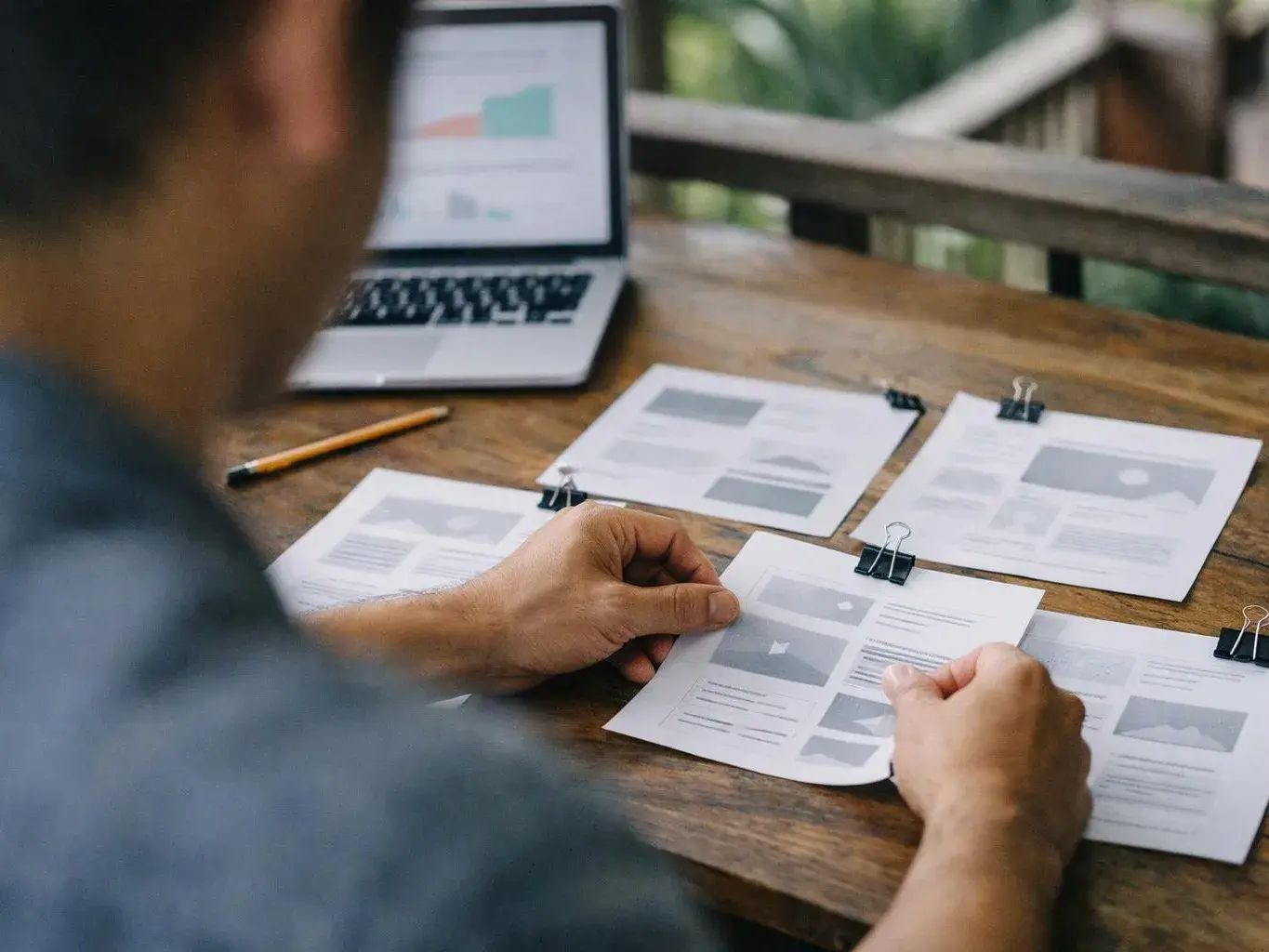 A person reviews clipped paper mockups beside a laptop on a wooden table outdoors