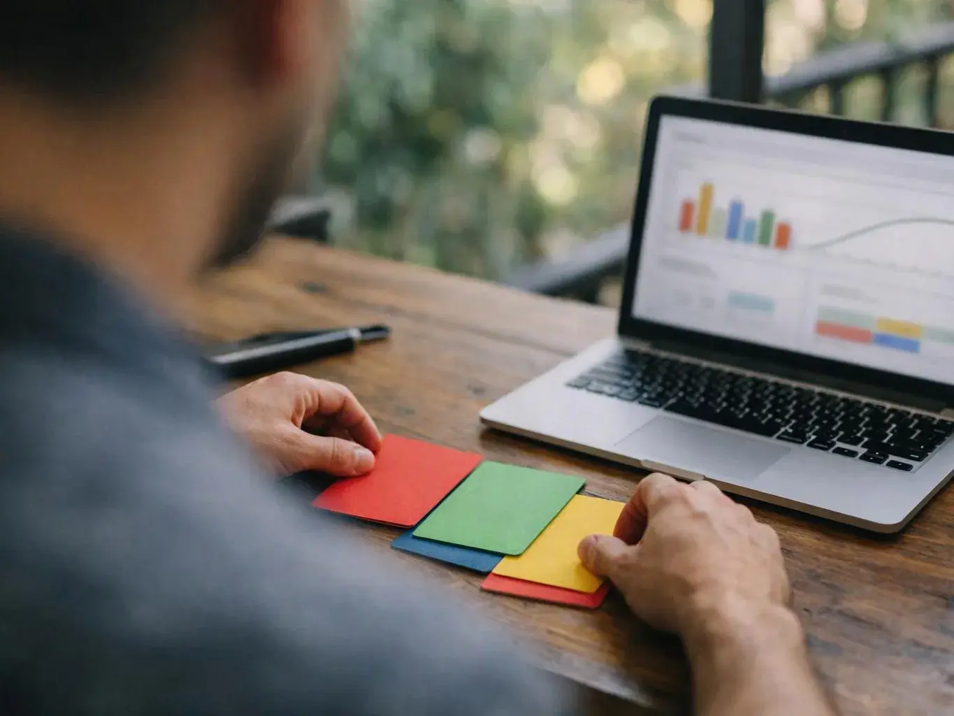 A person studies colourful sticky notes beside a laptop showing charts on a wooden table