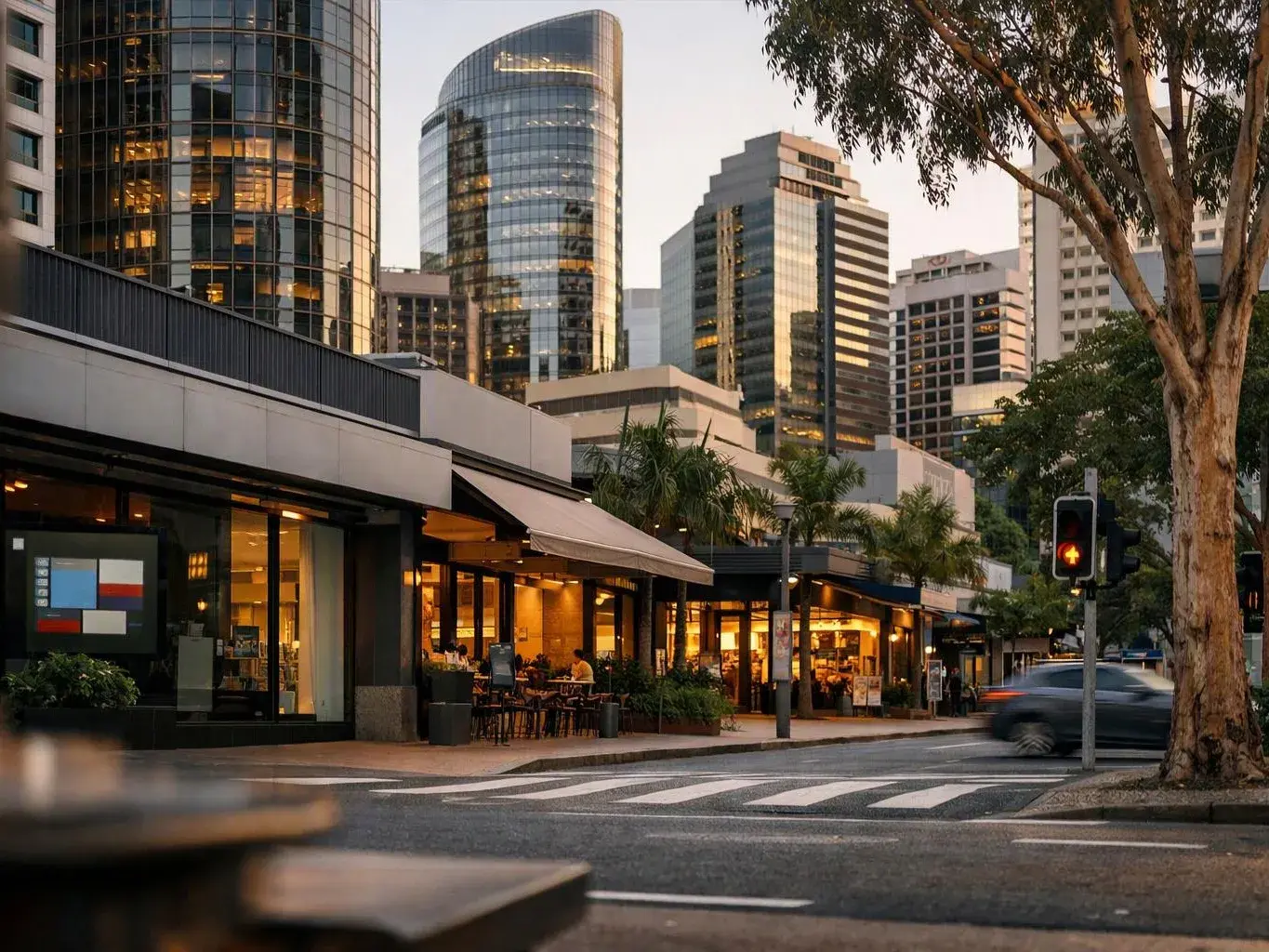 Sunset lights up glass towers above a street café and crosswalk with a passing car