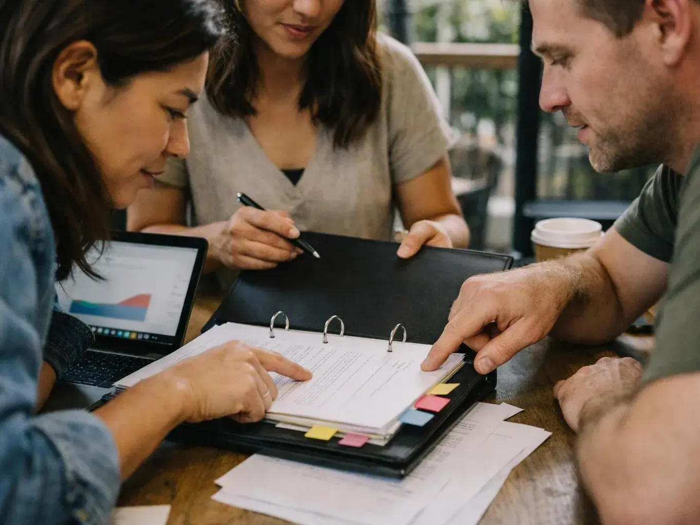 Three people review papers and a binder at a table, with a laptop and coffee cup nearby