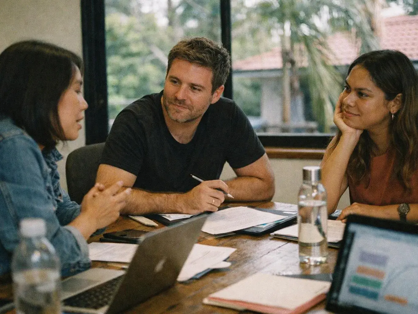Three people sit around a table, talking over papers, a laptop, and a water bottle in a bright room