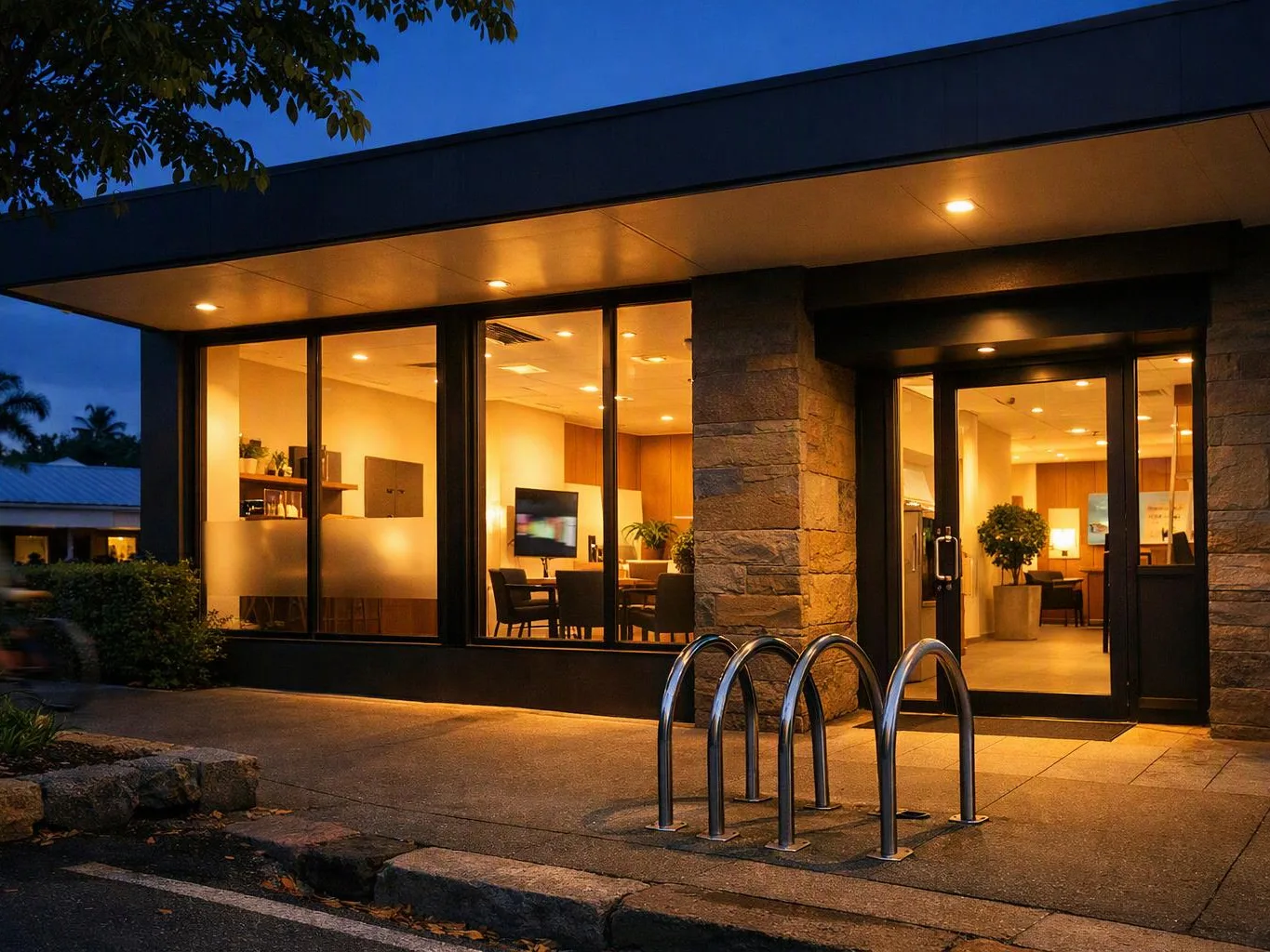 A warmly lit modern building entrance with large windows, glass doors, and a bike rack at dusk